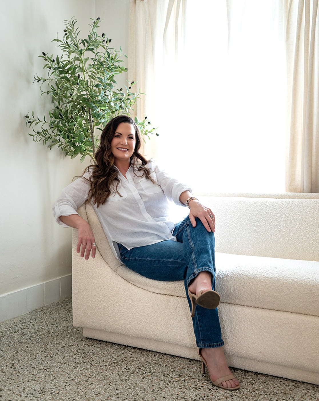 Woman sitting on a beige sofa in a bright room with a plant and window in the background